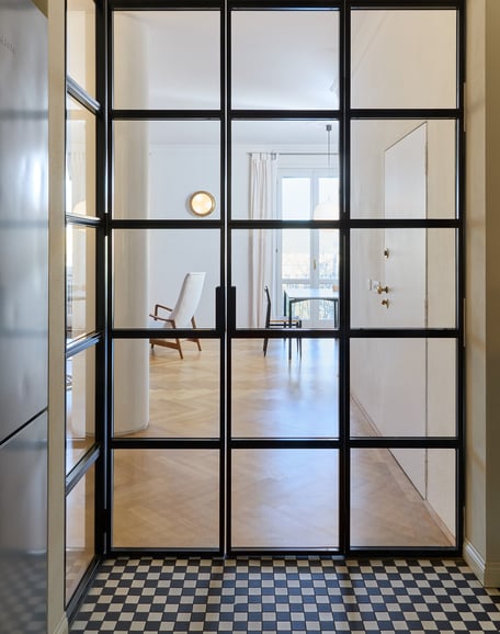 Vista del soggiorno dalla cucina, schermata da una porta scorrevole in vetro e ferro su disegno. Il parquet cede il passo a un pavimento in micro tessere black&white di grès, ricordo contemporaneo delle antiche marmette.