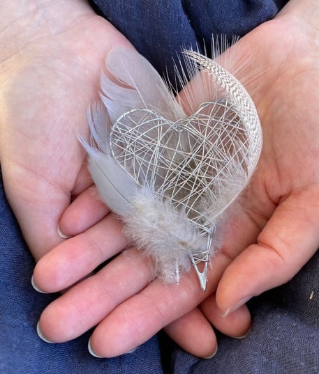 Zwei Hände halten ein silbernes Herz umgeben von weissen Federn.