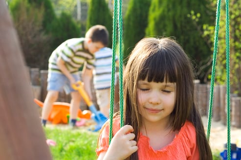 girl with her eyes closed on a swing, in the background boys playing with sand toys