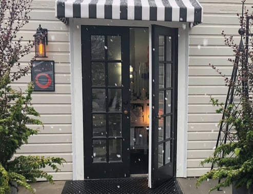 Photo showing the doors to STUDIO ORIGIN Intimate Garden Pilates Studio located in Newberg Oregon, in a chilly beautiful snow flurry. Black double doors, one slightly open to with a warm inviting soft lit glow inside.
