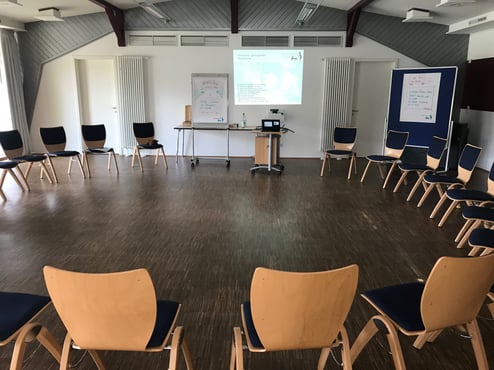 freundlich heller Semnarraum mit großem Stuhlkreis, Begrüßungsflipchart und bespannter Pinnwand