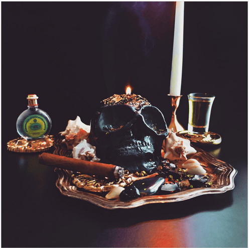 Ein zauberhafter schwarzer Kerzenschädel ruht auf einem verzierten Teller, umgeben von Kerzen, Kräutern und einer kleinen Flasche 4711. Die Szene wird von Kerzenlicht beleuchtet.