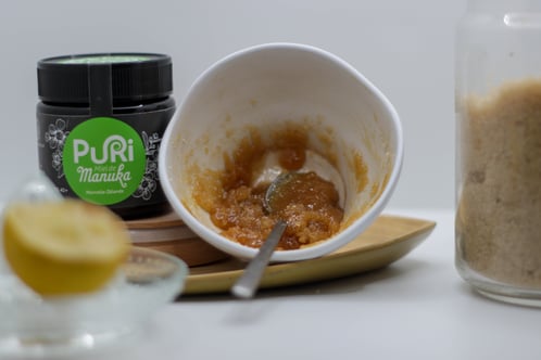 recette maison gommage au miel de manuka avec un pot de Miel de Manuka Puri New Zealand, un bol avec du miel du citron et de la cassonnade mélangé, cassonnade dans pot en verre et zest de citron au premier plan en fou le tout sous fond blanc