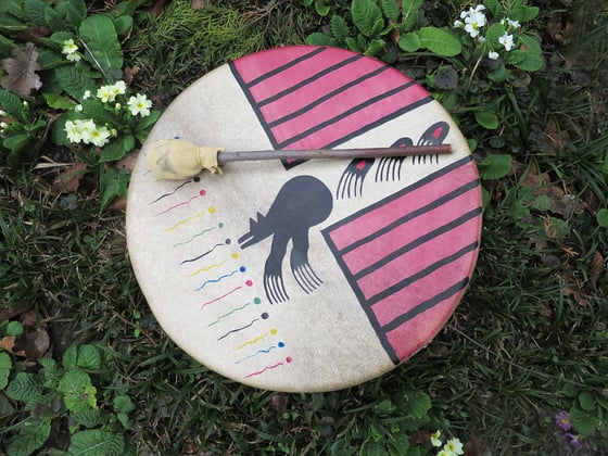 Eine schamanische Trommel mit schläger die auf dem Gras liegt umgeben von einzelnen blumen. Auf der Trommel ist der Bär zu sehen