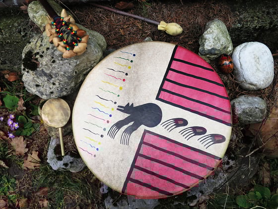 Eine grosse schamanische Trommel mit einem Bären und Tazen in Rot und schwarz abgebildet daneben liegt der Schlägel Rasseln und kleine Trommel und Rasselei liegen daneben auf dem Boden neben Steinen und Blumen