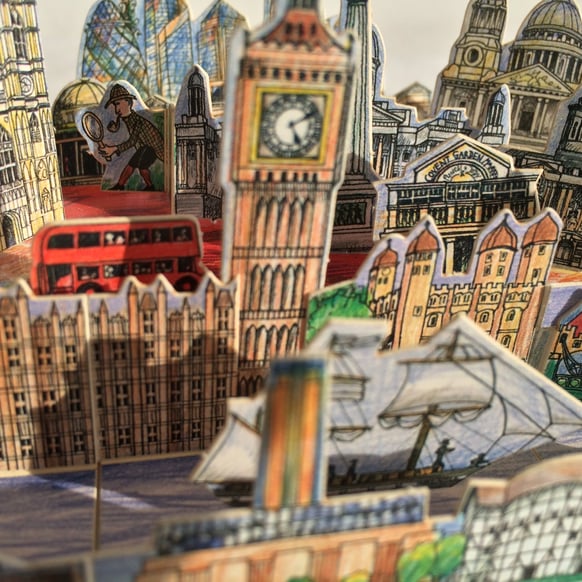 ein dreidmensionales Papiermodell von London mit Westminster, Big Ben, dem Tower und weiteren Gebäuden. Zwischen ihnen fährt ein Doppeldecker-Bus und im Hintergrund untersucht Sherlock Holmes etwas. Die Teile passen in eine Streichholzschachtel.