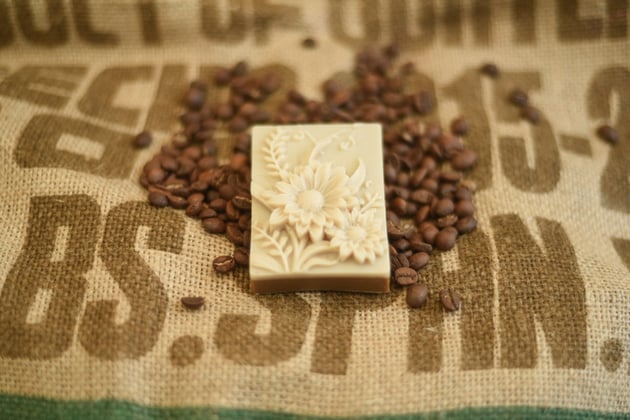 Das Bild zeigt eine Kaffeeseife mit Blumen auf Kaffeebohnen von Schneider Kaffee sowie einem Jutesack.