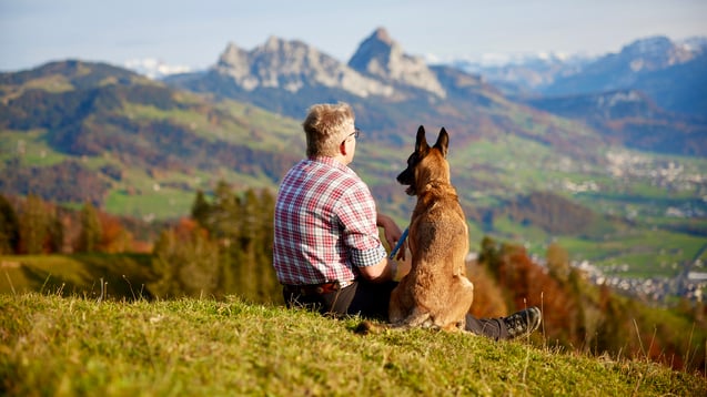 Ronald Kalbermatten und sein Hund Blue sitzen entspannt vor einer Alpwiese, Ronald schaut freundlich in die Ferne.