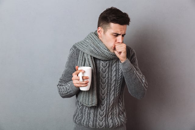 Homme avec un pull en laine pour l'hiver et une grosse écharpe autour du coup, il tousse et est malade. Dans sa main un mug avec une infusion