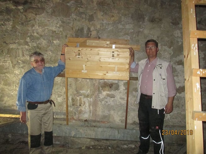 Zwei Männer in Arbeitskleidung stehen an einem großen Holzkasten an der Mauer
