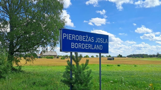 BNluse sign announcing "borderland" in Lativan and English in Latgale, Latvia, with a tree, a farmhouse, green fields and blue sky in the background