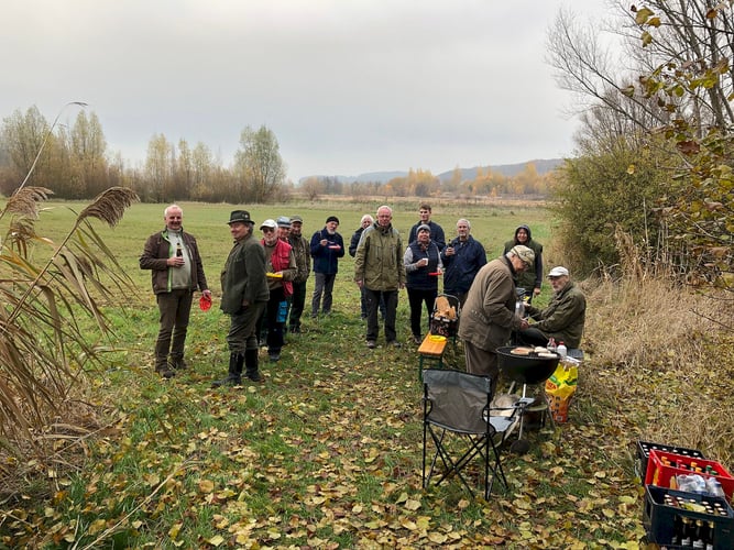 Eine Gruppe von 14 Personen vor einer Wiese und Hecke mit Grillwürstchen und Getränken
