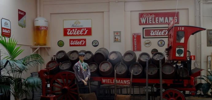 Photo: Le vieux camion rouge de Wielemans chargé de tonneaux devant un décor mural fait de panneaux publicitaires Wiels et Wielemans et d’un énorme verre de bière en hauteur dans le coin. Devant le camion, le mannequin d’un livreur.
