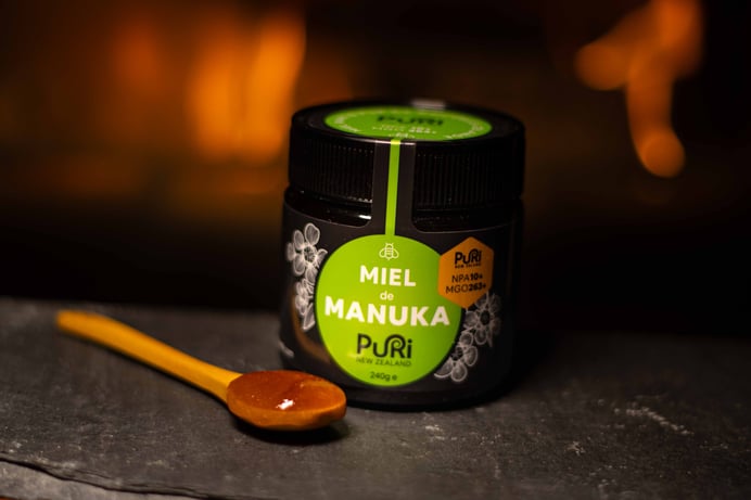 Miel de Manuka NPA 10+ / MGO 263+ Puri New Zealand en pot en verre de 240g et sa cuillère en bois avec du Miel de Manuka dedans sur une ardoise sous fond avec un feu d'un poêle avec des flammes chaleureuses