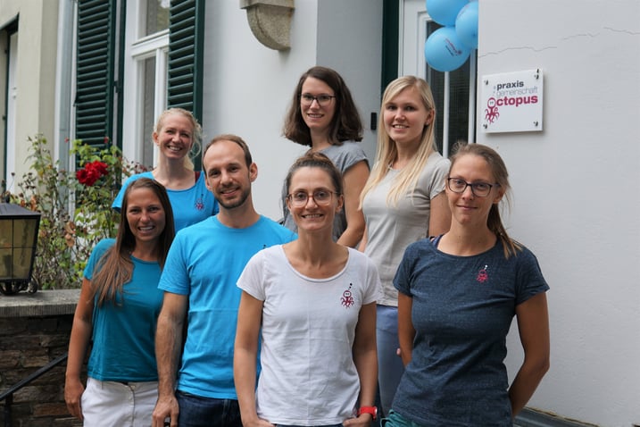 Das Therapeuten-Team der Praxis steht vor dem Eingang des Gebäudes. Alle haben ein Arbeitsshirt mit dem Praxislogo an, in verschiedenen blauen und grauen Farbtönen. Im Hintergrund sind Blumen, Luftballons und das Praxisschild zu sehen.