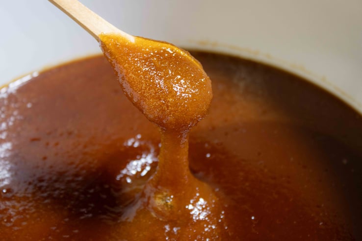 Une cuillère en bois couverte de Miel de Manuka NPA 24+ / MGO 1122+ de Nouvelle-Zélande dégoulinant dans un seau de 10kg, le Miel est orangé sombre et est épais