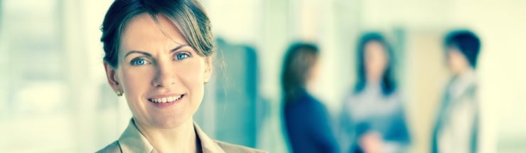 32jährige Frau mit dunkleren Haaren die sie im Nacken zusammen gebunden trägt. Hat blaue Augen und lächelt. Sie trägt eine beigen Blazer und im Hintergrund stehen 3 weitere "Business-Frauen" und sprechen miteinander.