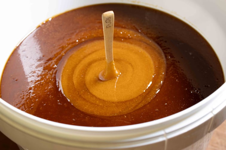 Cuillère en bois Puri New Zealand plantée dans un seau de 10kg de Miel de Manuka NPA 24+ / MGO 1122+ le centre du miel est clair et mélangé, les bords extérieurs plus sombres