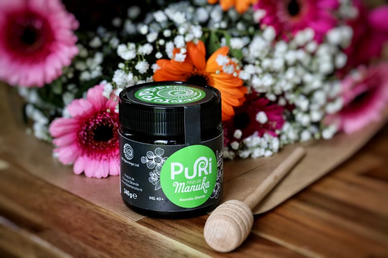 Pot en verre 240g de Miel de Manuka Puri New Zealand avec une cuillère en bois sur une table en bois avec un magnifique bouquet de fleurs en arrière-plan avec des fleurs roses, oranges et blanches