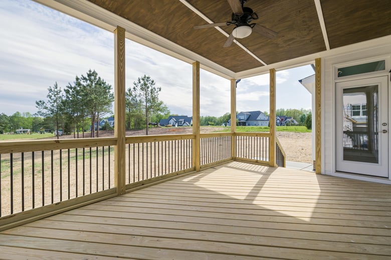 A spacious, covered wooden deck with a ceiling fan overlooks an open yard with trees and houses in the distance, offering a serene and relaxing outdoor setting.