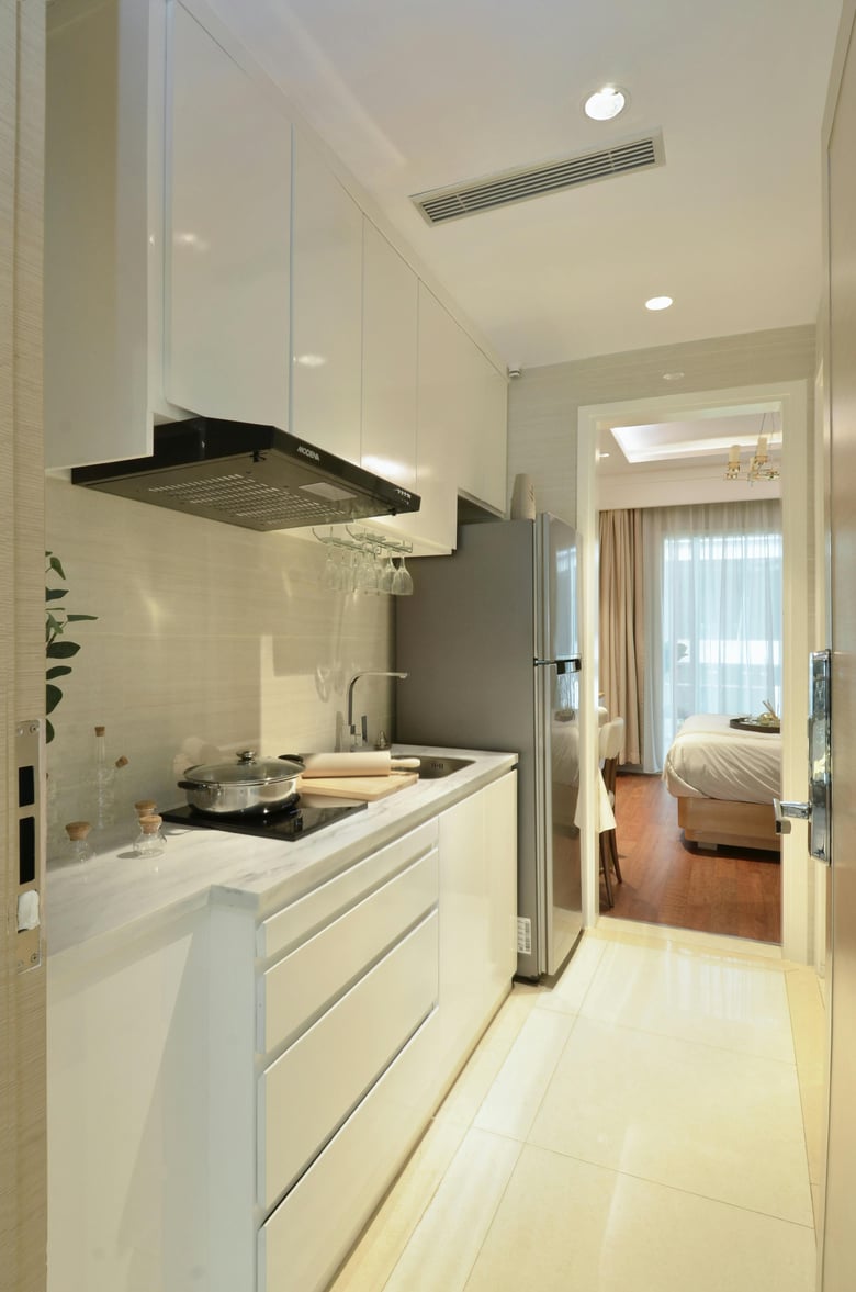 A compact and modern kitchen space with sleek white cabinets, integrated appliances, and a minimalist design, leading into a cozy bedroom with natural lighting.