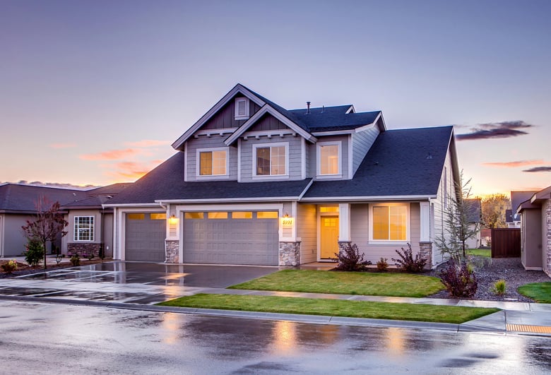 A modern blue and gray concrete house with an attic and a double-door garage stands against the twilight sky, its soft glow reflecting off the neatly manicured green grass lawn in the front.