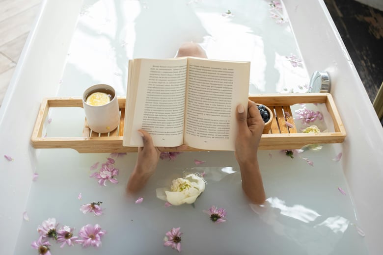 A person relaxes in a floral bath, reading a book with a cup of tea and a bowl of berries on a wooden bath tray.