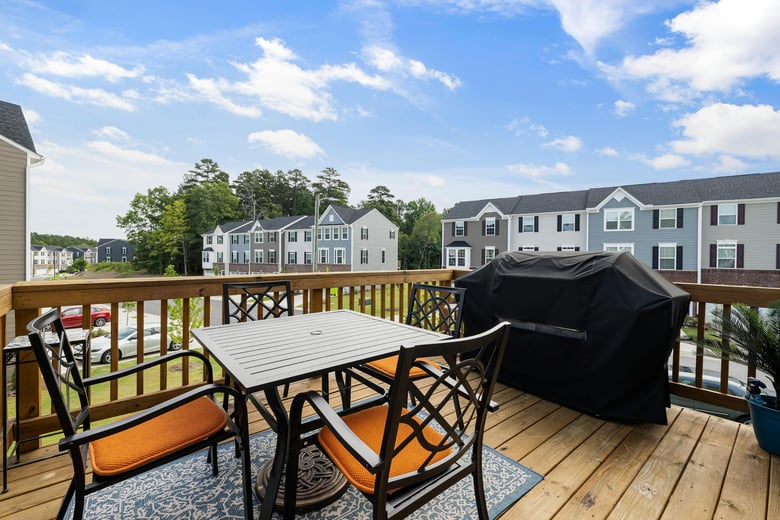 A cozy outdoor deck features a modern black dining set with orange cushions, a covered grill, and a view of neighboring townhouses under a bright blue sky.