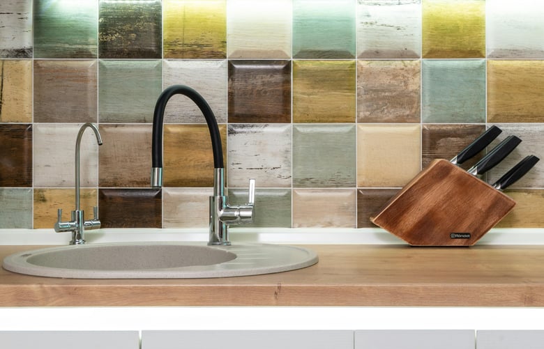 This kitchen setup features a sleek sink with a black and chrome faucet, a wooden countertop, a colorful tiled backsplash, and a wooden knife block, blending natural and modern elements