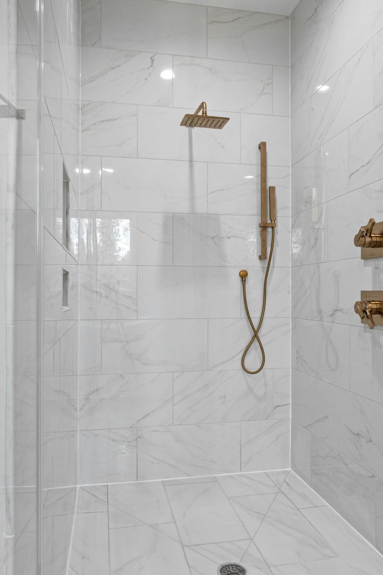 a sleek, modern shower with large white marble tiles and gold fixtures, including both a rainfall showerhead and a handheld attachment.