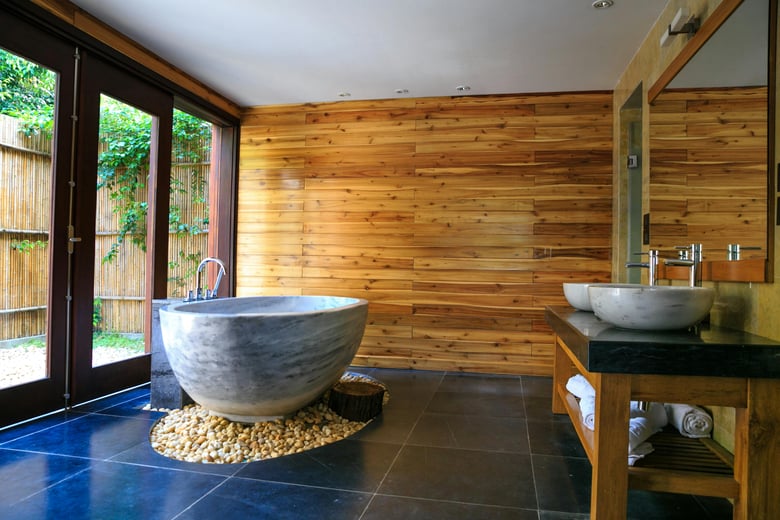 This bathroom exudes a serene spa-like atmosphere with its large stone bathtub set on pebbles, warm wooden walls, and natural light streaming in from large glass doors.