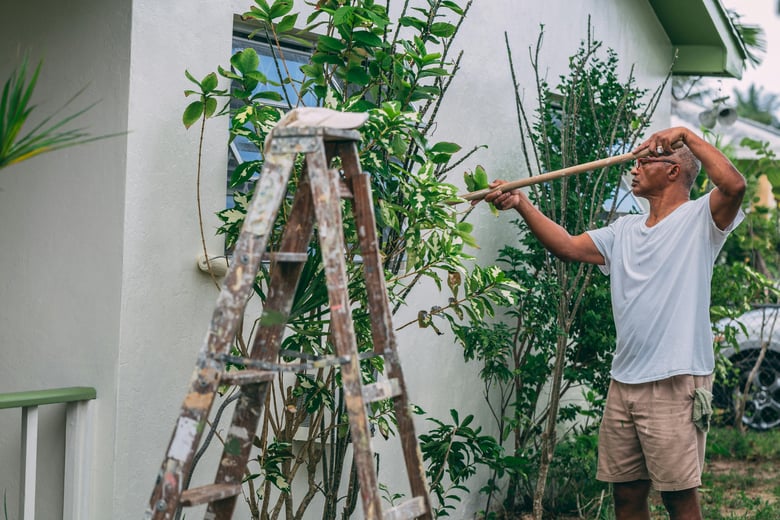 An house owner in casual clothes is trimming plants with a long-handled tool in a garden next to a white house, while a paint-splattered ladder stands nearby