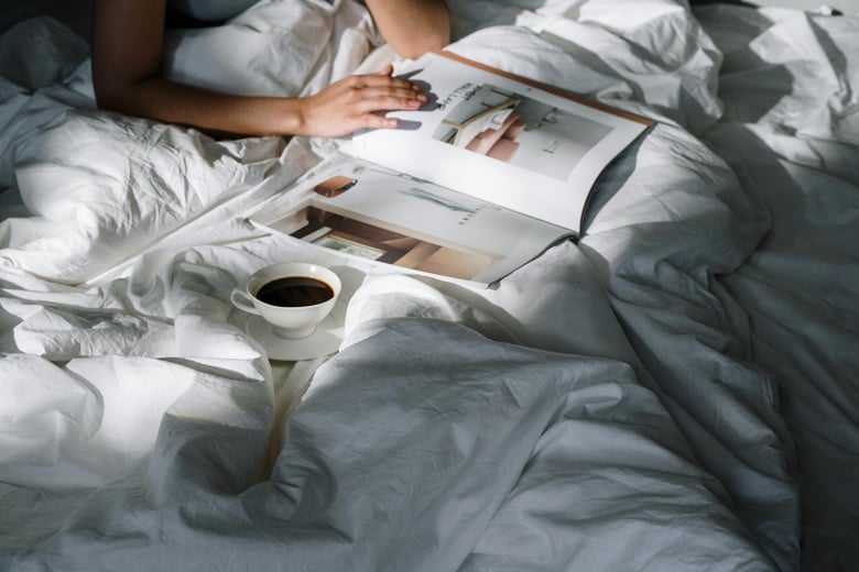 A serene morning moment captured, with a person enjoying a magazine and a cup of coffee amidst soft, crumpled white bedding.
