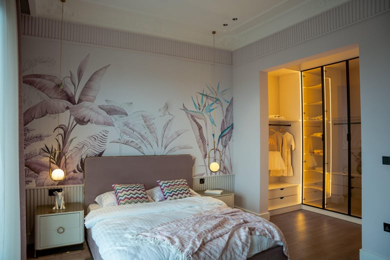 A modern bedroom with grey bed linen, two white side tables with pendant lights, and a walk-in wardrobe with glass doors, LED lighting, and laminated floating floorboards.