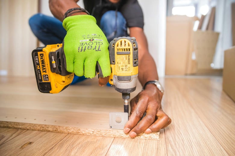 A kitchen contractor, equipped with safety gear, is assembling a kitchen kickboard using a DeWalt electric drill