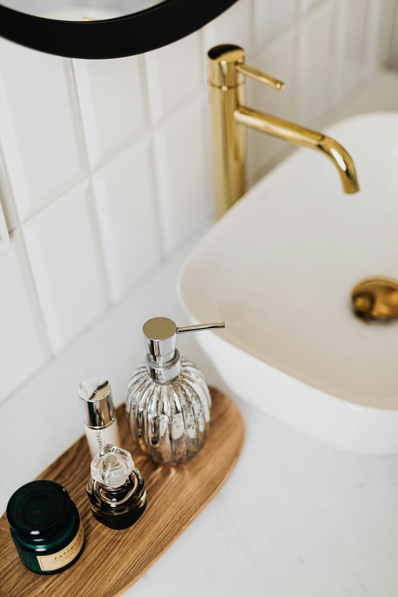 A stylish bathroom sink area features gold fixtures and a wooden tray holding a collection of elegant toiletries and a silver soap dispenser.