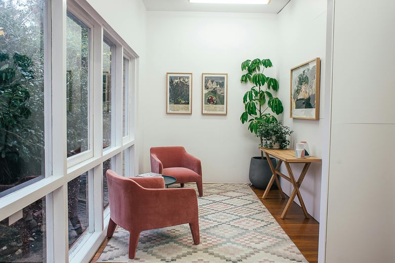 This is a cozy and minimalist corner with soft pink chairs, a patterned rug, potted plants, and framed artwork, framed by large windows that bring in natural light