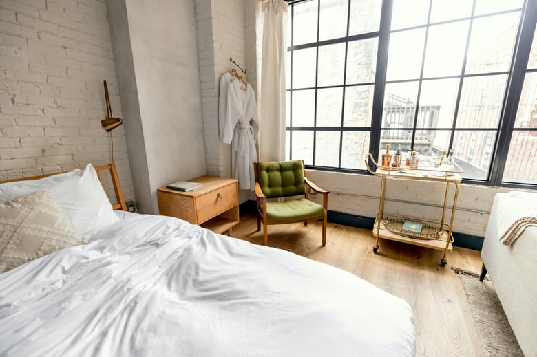A cozy bedroom features crisp white bed linen, a brown wooden chair, and a gold trolley stocked with whiskey, books, and glasses, all set against a stunning modern city view.