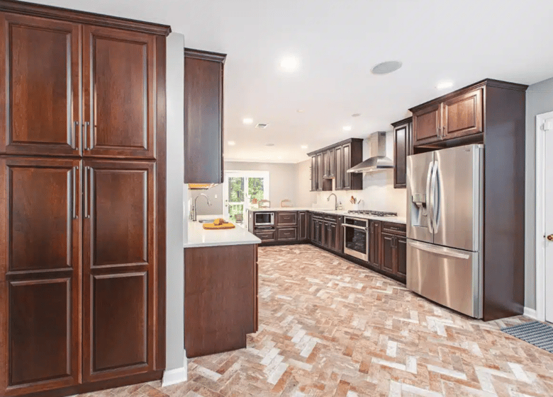 A spacious, modern kitchen featuring rich, dark wood cabinetry, stainless steel appliances, and a herringbone-patterned tile floor, all bathed in natural light.