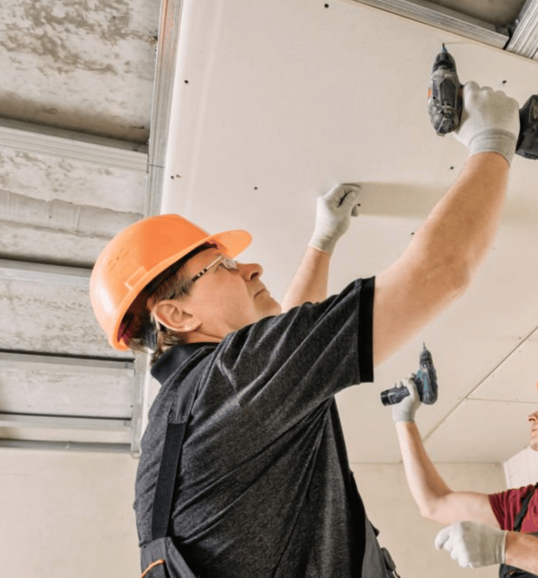 A construction worker wearing a hard hat uses a power drill to secure a ceiling panel in place.