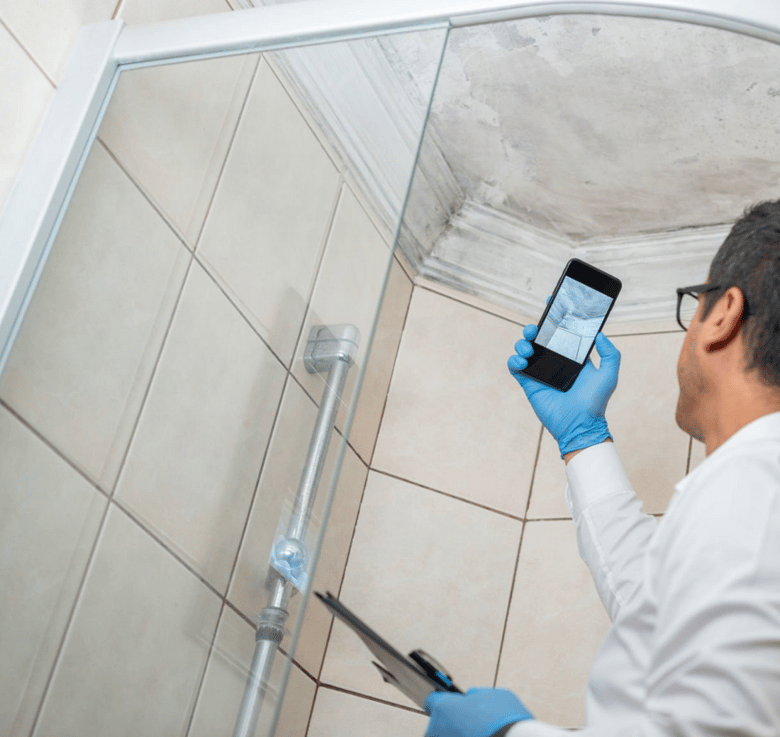 A person wearing gloves is inspecting and photographing mold growth on the ceiling corner of a tiled bathroom.