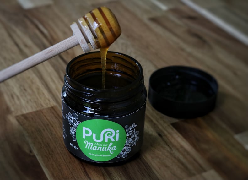 Miel de Manuka Puri New Zealand en pot en verre ambré de 240g sur une table en bois avec une cuillère à Miel qui laisse couler du Miel dans le pot