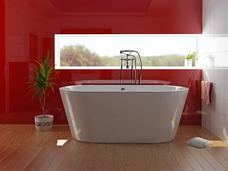 A bright, sunlit bathroom features a large white freestanding bathtub with chrome mixers, timber-look tiled floors, and a striking classic red glass feature wall that adds a bold touch of elegance.