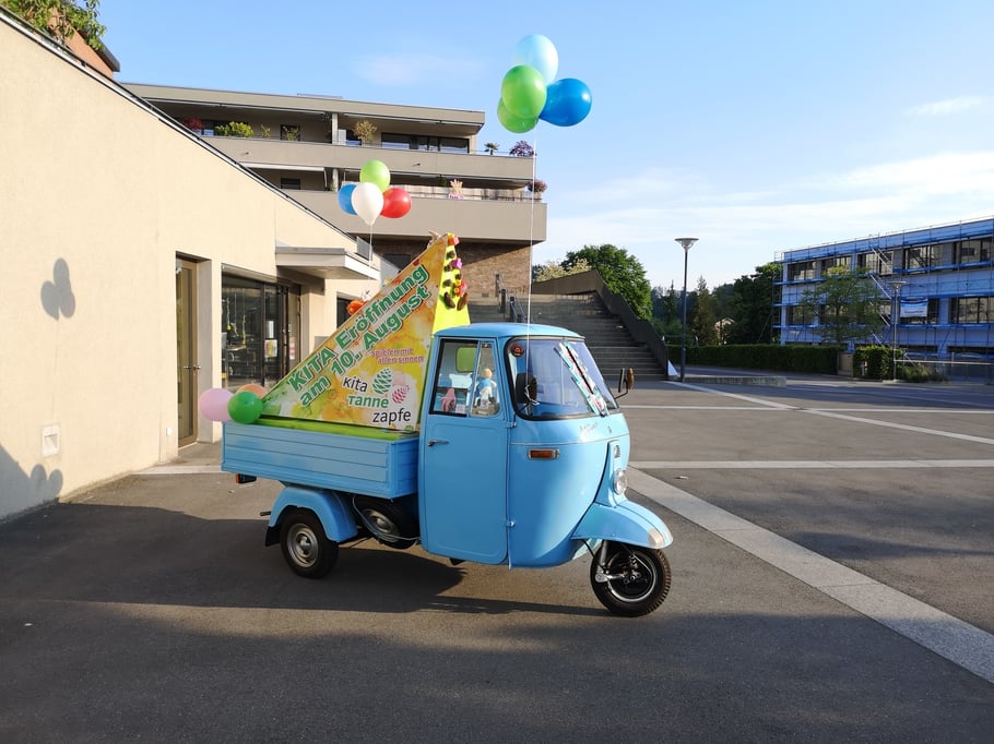 Nostalgisch 50-er Jahr blaues Piaggo-3-Rad mit Anhänger auf dem Dorfplatz von Langnau. Auf dem Anhänger als 3-eckiges Skulptur Werbung zur Kita-Eröffnung am 10. August.
