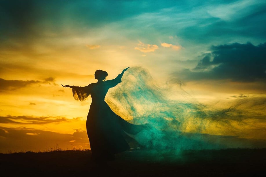 Silhouette einer Frau, die in einem langen Kleid tanzt, mit blauem und grünem Rauch auf dem Boden, vor einem Sonnenuntergangshimmel