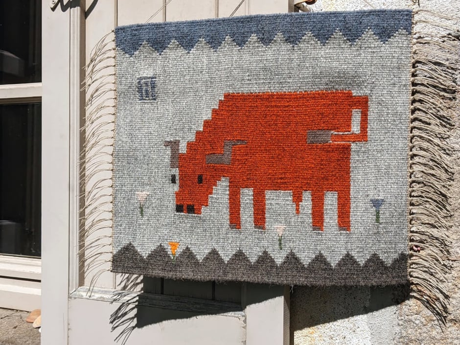 Un kilim-tenture murale en tissage représentant une vache salers qui regarde un papillon sur un pré de crocus. Dessin du tissage mural géométrisé.