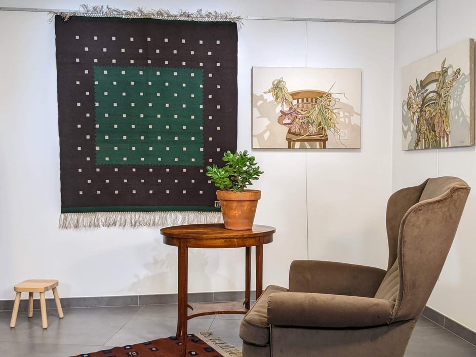 Intérieur avec, sur le mur, un kilim carré marron, motif carrés gris, tissé à la main en laine. À droite, une table avec une plante succulente en pot de terre cuite, devant un grand fauteuil voltaire et sur le mur au-dessus, deux tableaux - nature morte.