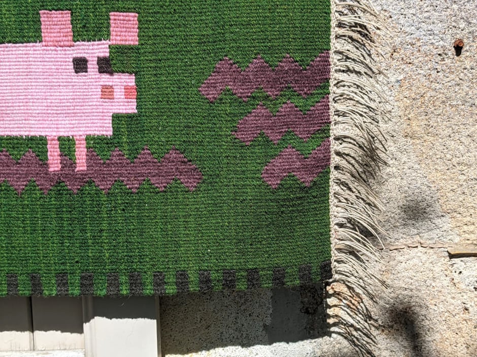Détail d'un kilim-tenture murale en tissage au fond vert et un petit cochon rose jouant dans la boue. Dessin du tissage mural géométrisé.