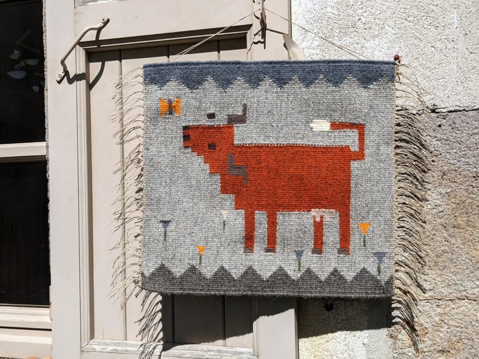 Un kilim-tenture murale en tissage représentant une vache salers qui regarde un papillon sur un pré de crocus. Dessin du tissage mural géométrisé.