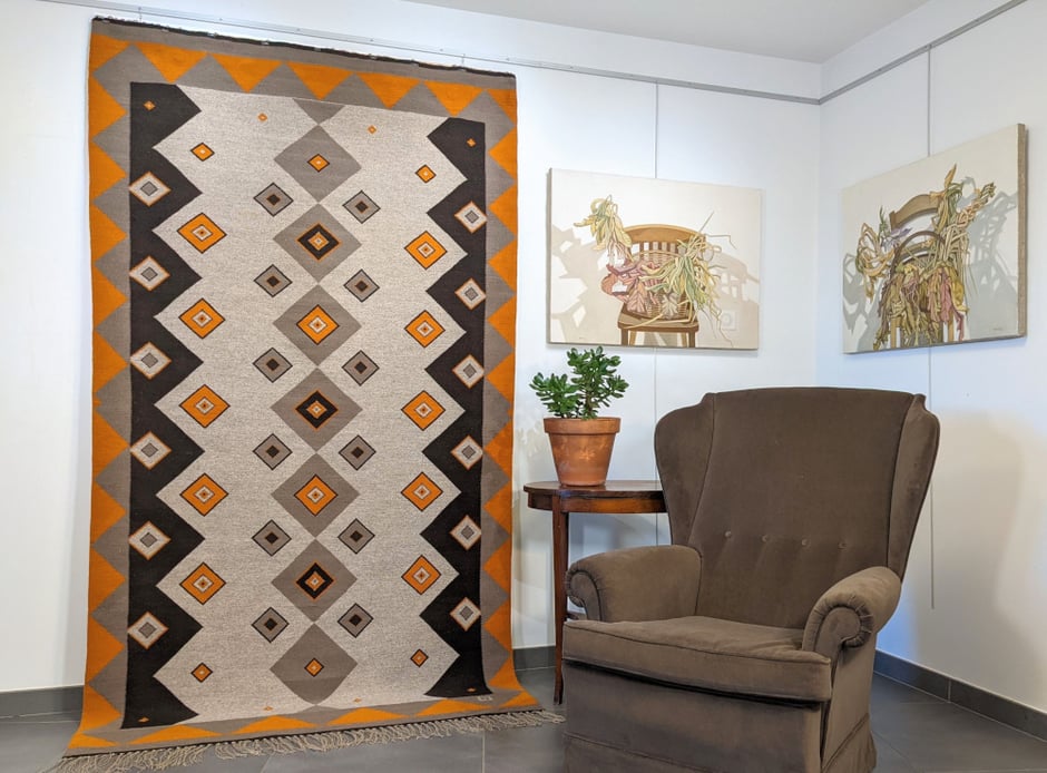 Intérieur avec, sur le mur, un grand kilim gris-orange-marron, tissé à la main en laine. À droite, une table avec une plante succulente en pot de terre cuite, devant un grand fauteuil voltaire et sur le mur au-dessus, deux tableaux - nature morte.